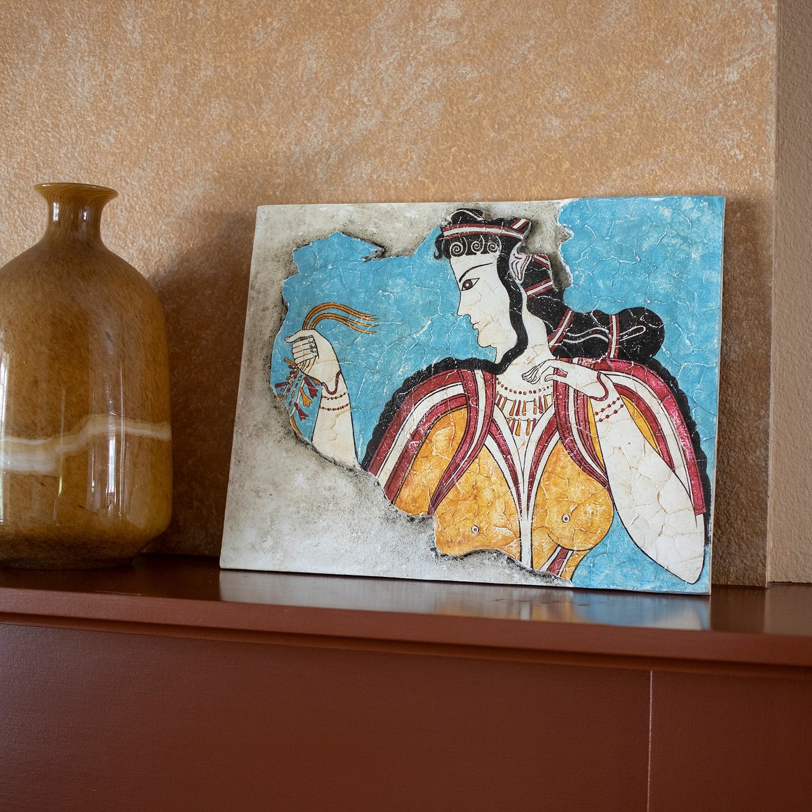Decorative tile with a woman's portrait and a brown vase on a shelf against a beige wall.
