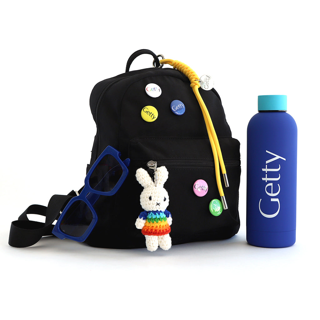 Image of black backpack next to the blue water bottle that says Getty. Backpack is decorated with a Miffy Rainbow Keychain, Rainbow Getty Buttons, and has blue sunglasses hanging off of it.