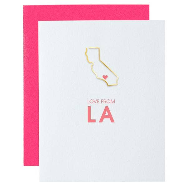 A white greeting card with a gold foil outline of California and a small pink heart, with the words 'LOVE FROM LA' in pink, shown with a bright pink envelope behind it on a white background.