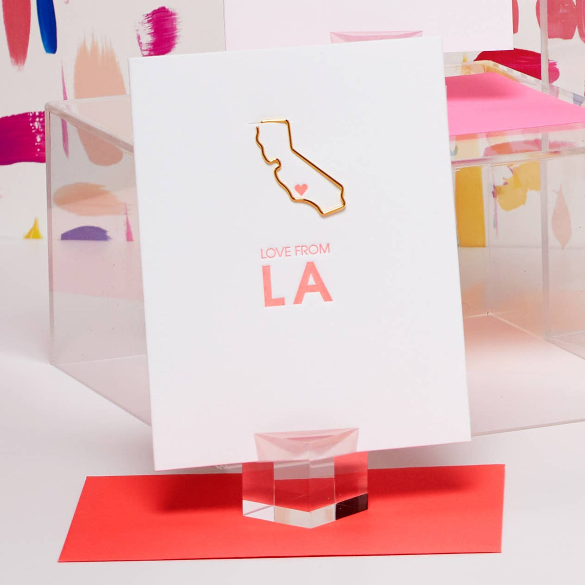 A white greeting card with a gold foil outline of California and a small pink heart, plus the words 'LOVE FROM LA' in pink, displayed on a clear acrylic stand over a red card on a white surface.