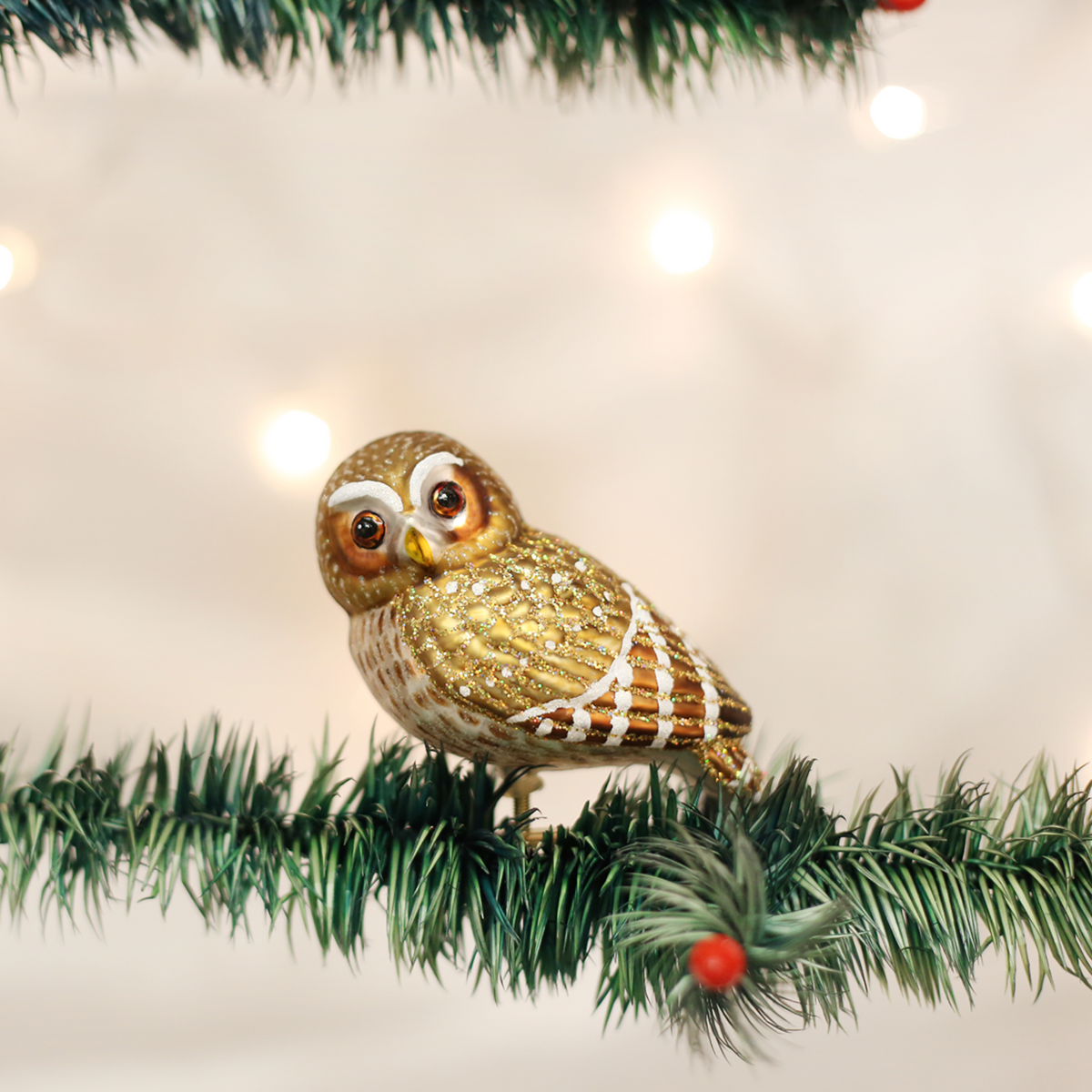A small decorative owl ornament perched on a green artificial Christmas tree branch, with warm blurred lights in the background.