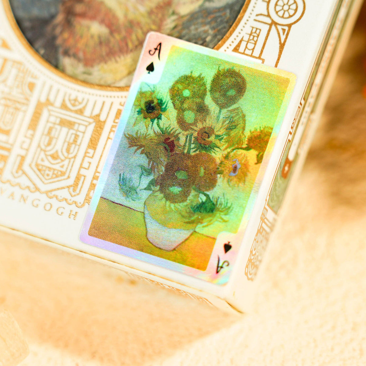A close-up of a Van Gogh-themed playing card and its decorative box, featuring a sunflower painting design with a holographic shimmer effect, set against a warm beige background with blurred red flowers.
