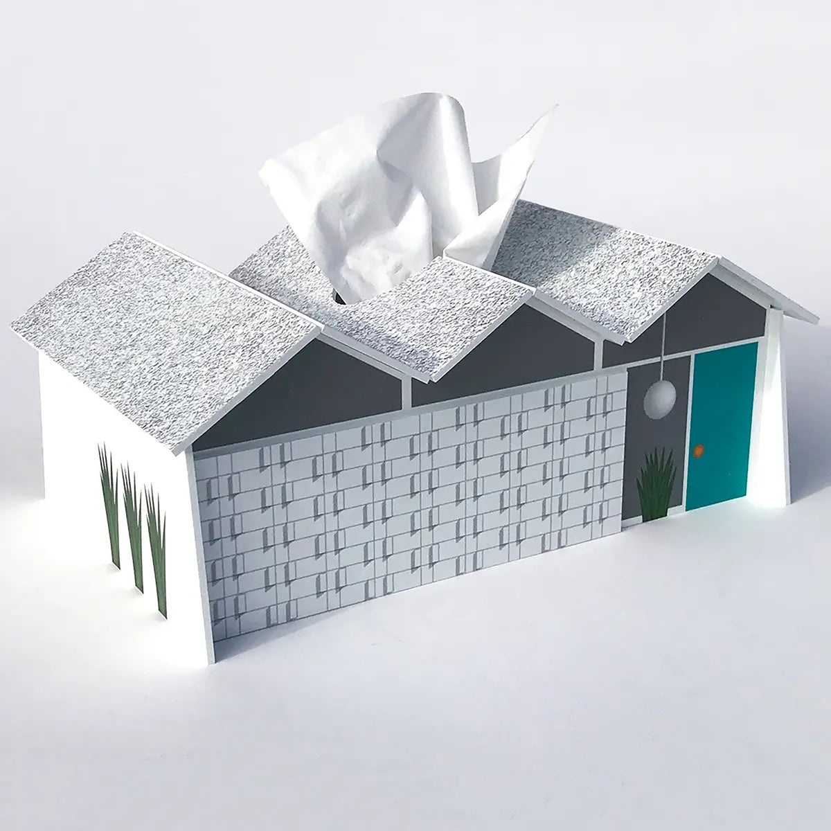 A novelty tissue box cover shaped like a house, with a white tissue protruding from the roof opening. The house design has a textured silver-gray roof, a white shingled wall, dark gray side panels, a teal door, and small green plant details, shown on a clean white background.