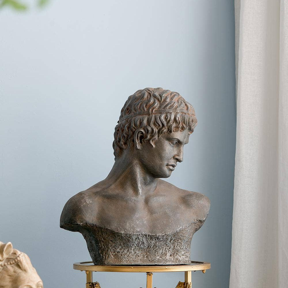 A classical-style bronze bust sculpture of a young man with curly hair, displayed on a gold metal stand against a light blue wall with a white curtain.