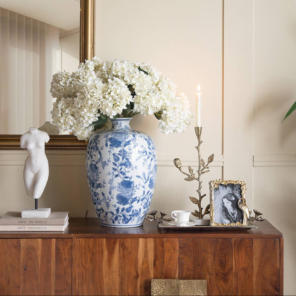 A blue and white porcelain vase filled with white hydrangeas sits on a wooden console table in a styled interior setting with a gold mirror, white sculpture, and candle holder.