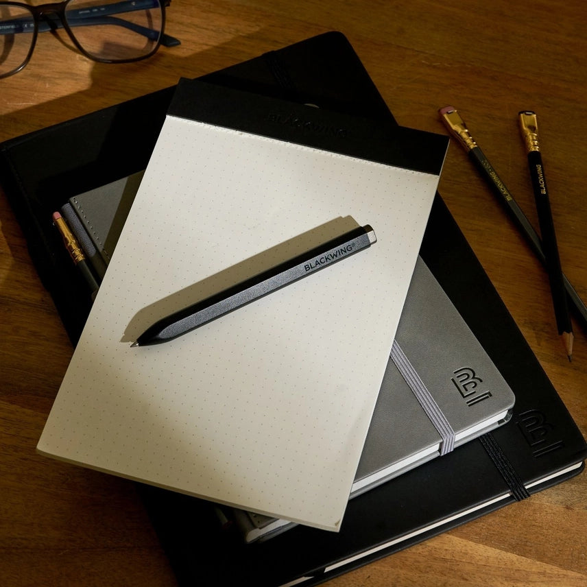 Notebooks and a pen on a wooden surface with glasses and pencils in the background