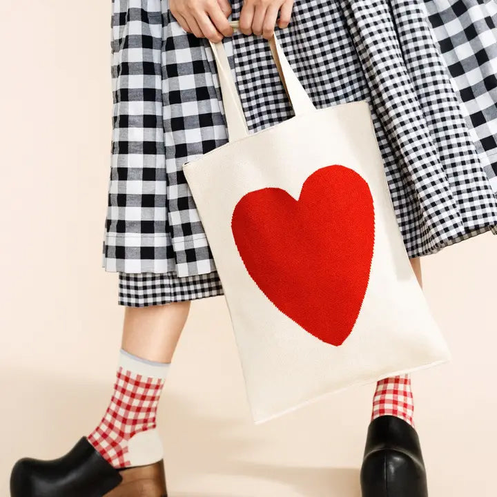 Person holding a tote bag with a red heart design, wearing checkered pants and socks.