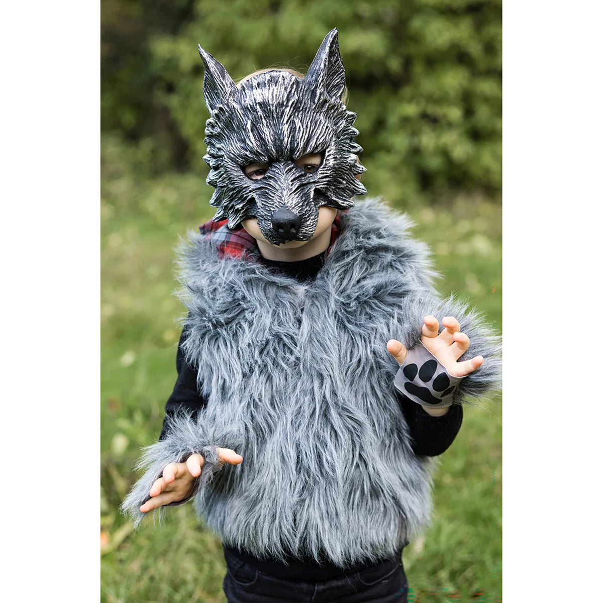 A child wearing a gray faux-fur wolf costume vest with matching paw-print wristbands and a detailed gray wolf mask, standing outdoors in a grassy field.