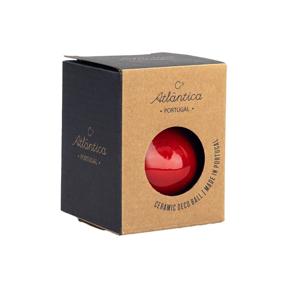 A red glossy ceramic decorative ball visible through a circular cutout on a brown kraft paper box with black sides. The box is branded Atlantica Portugal.