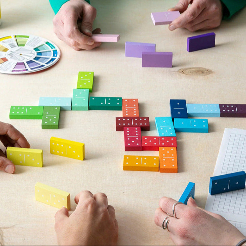 A group of people's hands arranging colorful wooden domino blocks on a light wood tabletop. The set includes dominoes in many bright colors such as green, blue, yellow, orange, red, pink, and purple, along with a color wheel and a graph paper sheet.