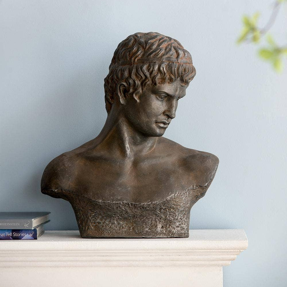 A classical-style bronze bust sculpture of a young man's head and upper chest, displayed on a white mantelpiece against a pale blue wall with soft green leaves in the background.