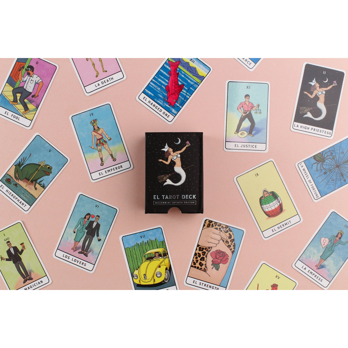 El Tarot Deck box stands upright in the center of a pink surface, surrounded by scattered illustrated tarot cards. The box is black with white text and a white mermaid illustration, while the cards around it show colorful tarot imagery on pastel and bright backgrounds.