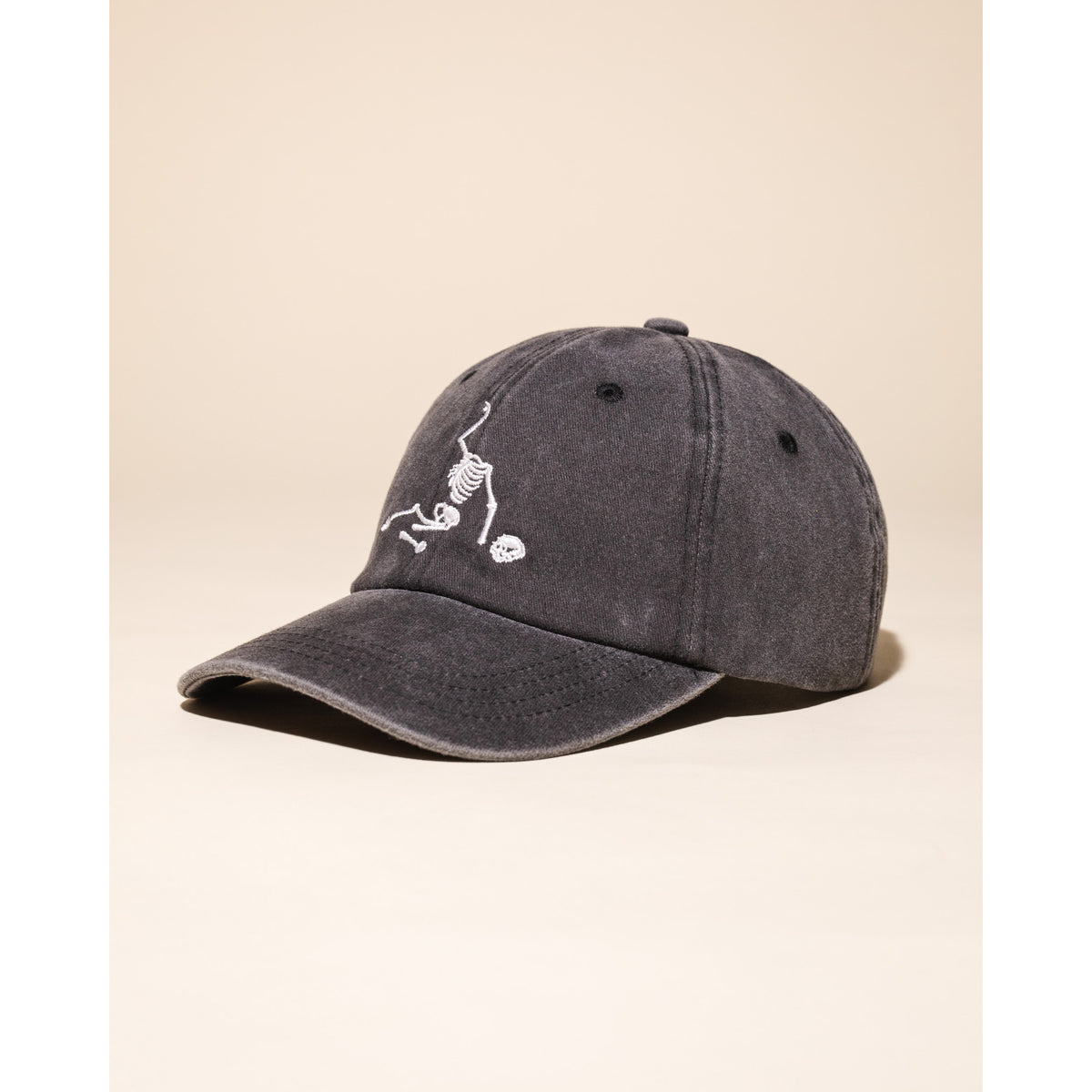 A dark gray washed baseball cap featuring a white embroidered skeleton holding a skull on the front, set against a solid beige background.