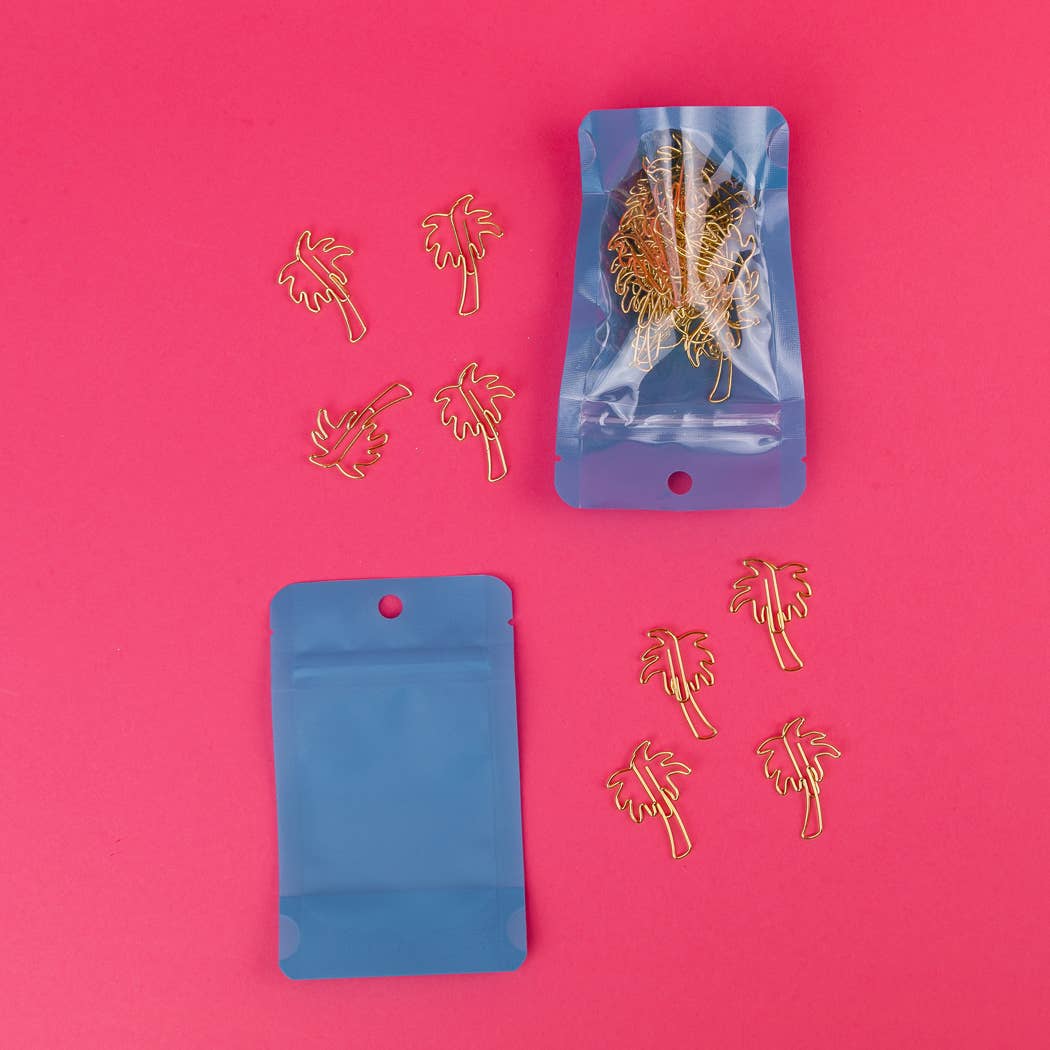 Gold metal palm tree wire hooks displayed on a bright pink background, with two blue zip-top pouches shown alongside one filled with the hooks and one empty.