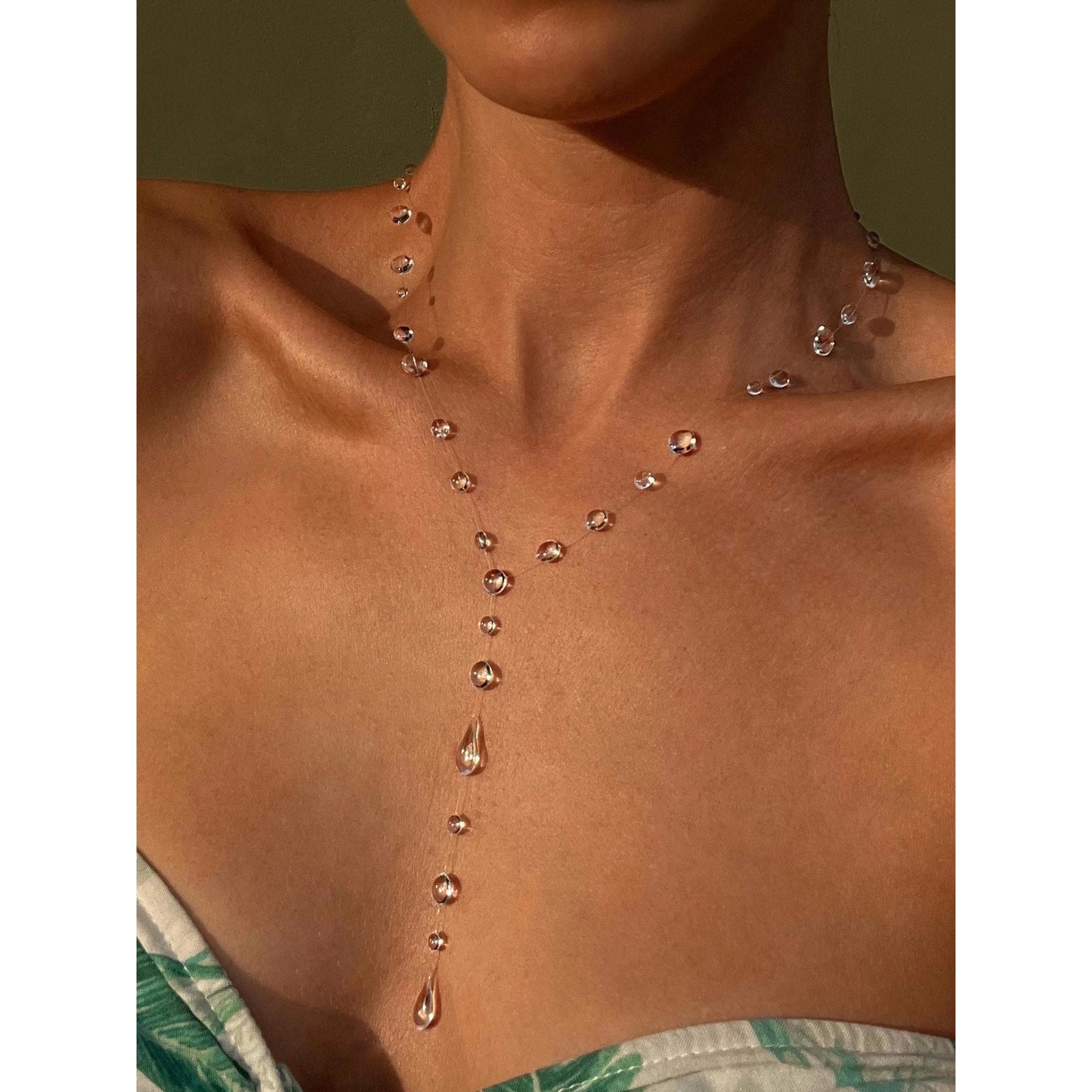 Close-up of a person with a necklace that resembles water droplets on their skin against a neutral background