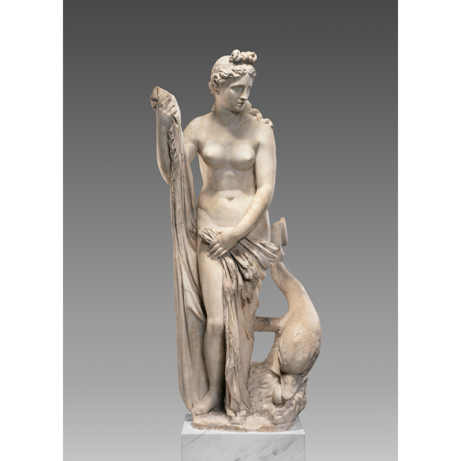 A white pin shaped like a statue of Venus, goddess of love, standing nude and holding a cloth at her hips, with a dolphin at her feet.