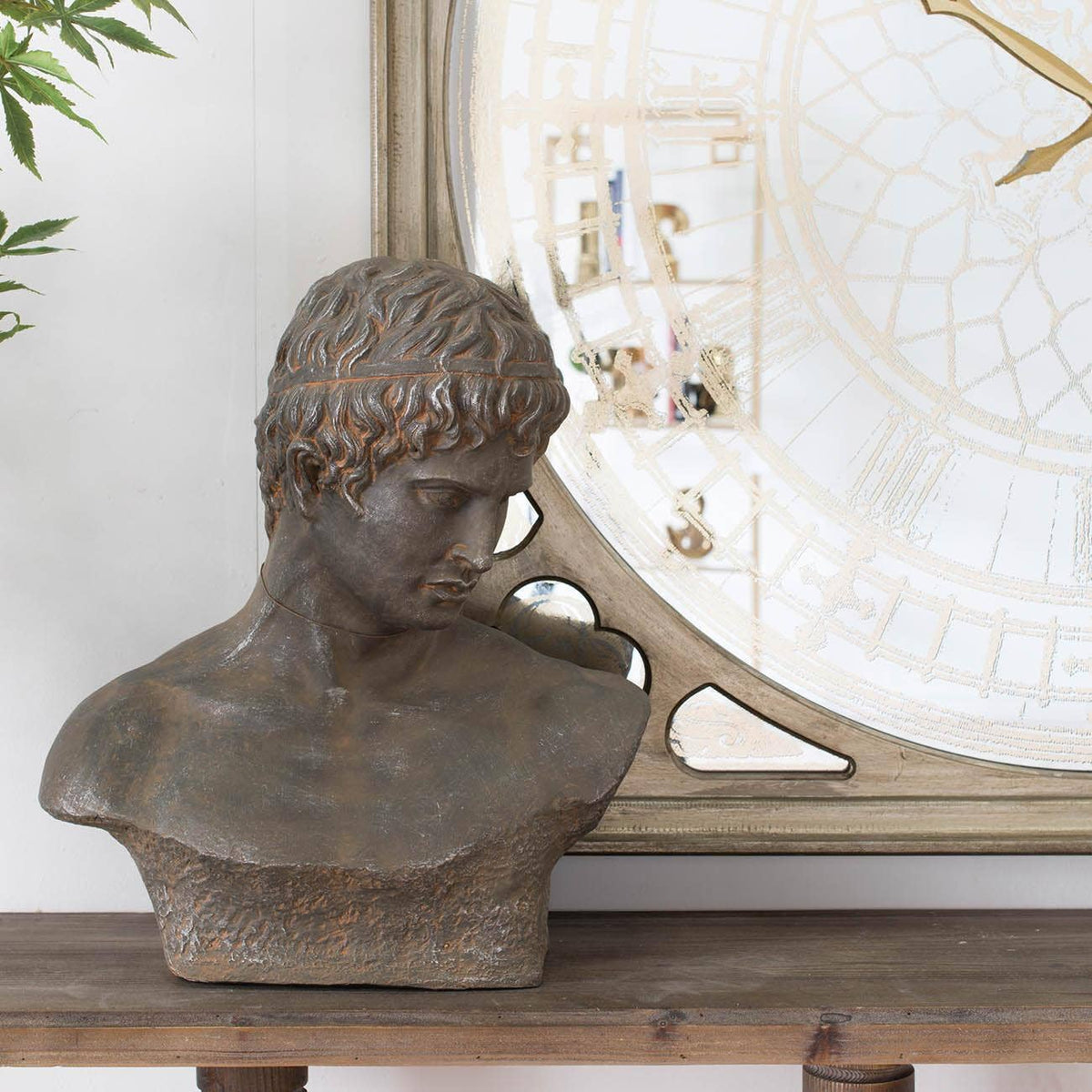 A bronze-finished classical male bust sculpture displayed on a wooden console table in front of a large decorative mirror.