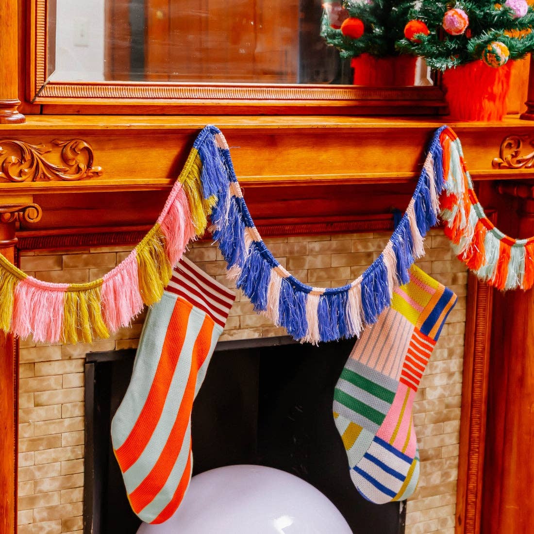 Three festive Christmas stockings with colorful striped and patchwork patterns hang from a wooden mantel decorated with a pink, blue, and yellow fringe garland. The fireplace has a beige brick surround, and a small decorated Christmas tree is visible in the background.