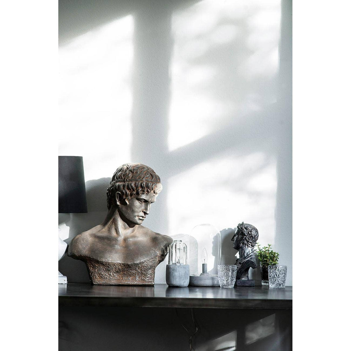 A decorative classical-style bust sculpture in a weathered bronze finish sits on a dark console table in a modern interior. Strong diagonal sunlight casts shadows across a light gray wall behind it, alongside a smaller dark figurine and small plants.
