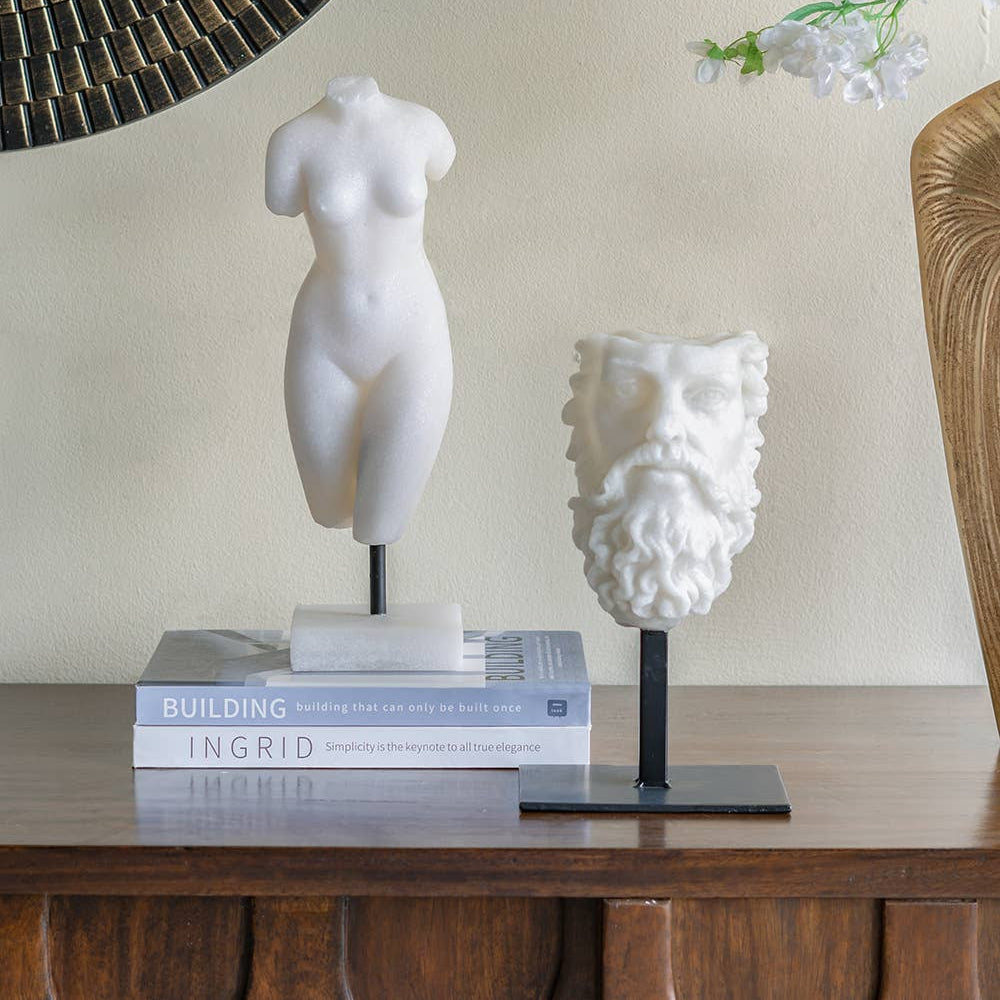 Decorative items including a marble sculpture, a bust, and books on a wooden surface with a mirror and flowers in the background.