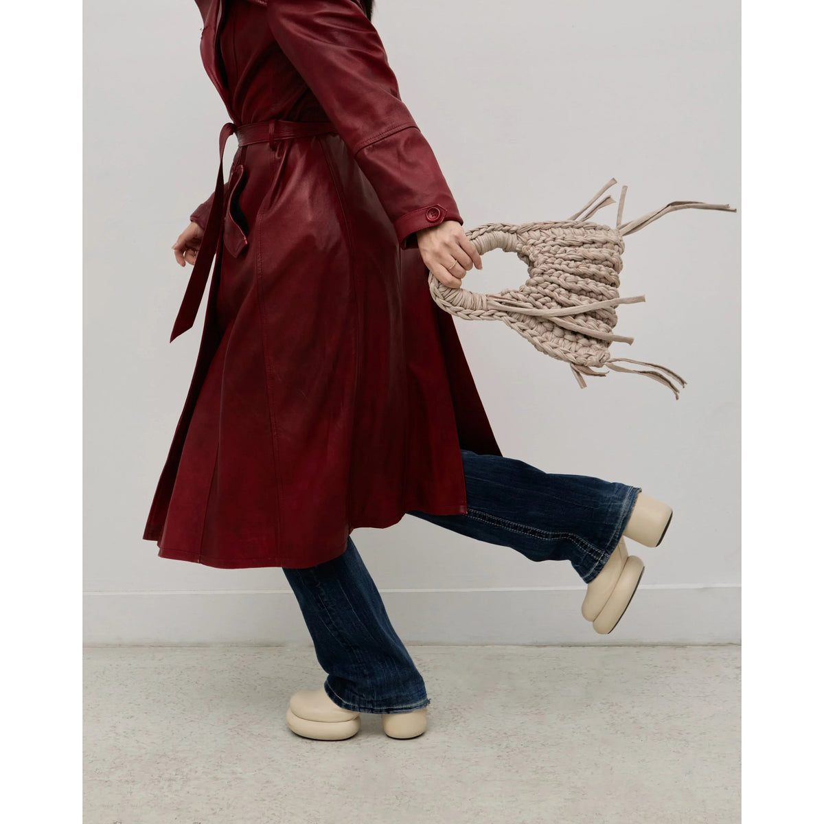 Person wearing a red coat and blue jeans, holding a beige bag against a white background