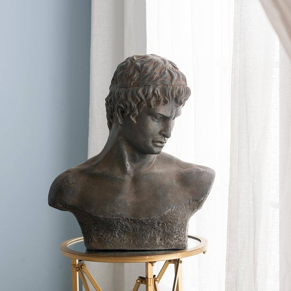 A classical-style bronze bust sculpture of a young man, displayed on a gold metal side table in front of white sheer curtains and a light blue wall.