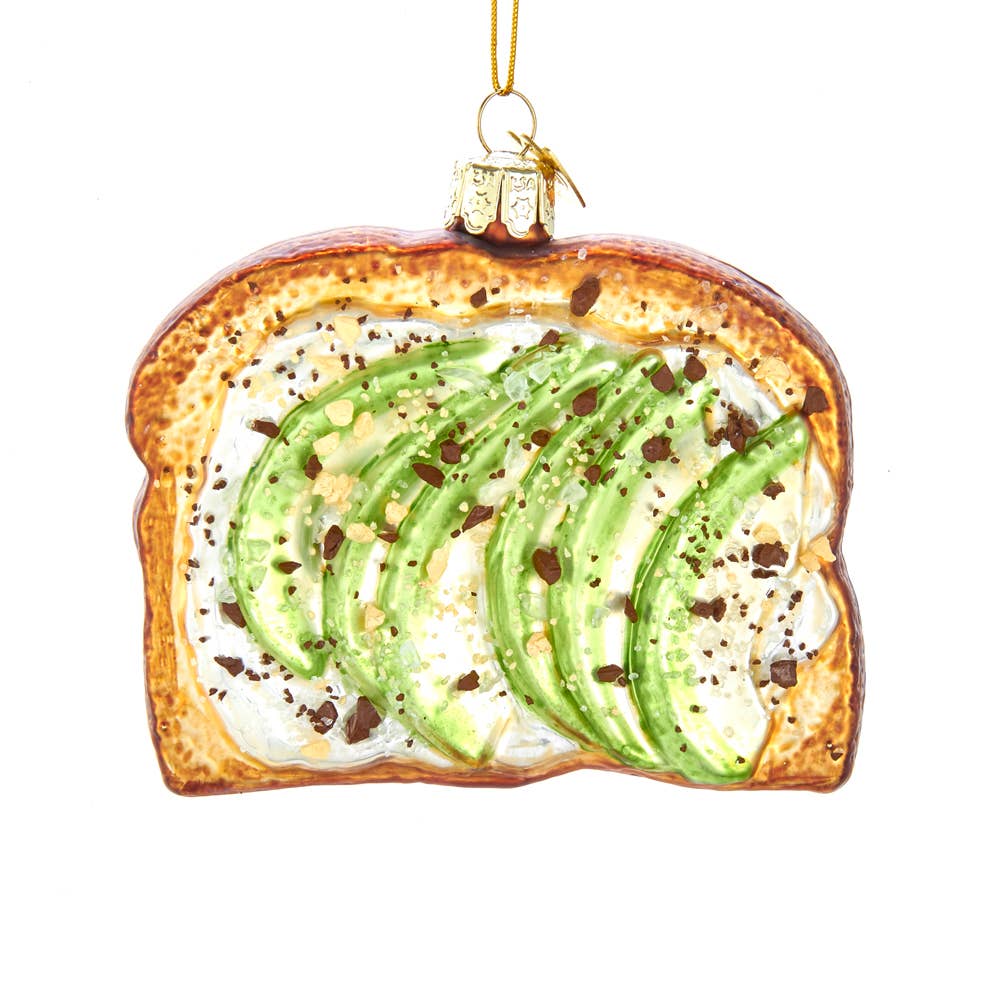 A glass Christmas ornament shaped like a slice of avocado toast, featuring green avocado slices on a white spread background with brown toast edges, hanging from a gold string against a white background.