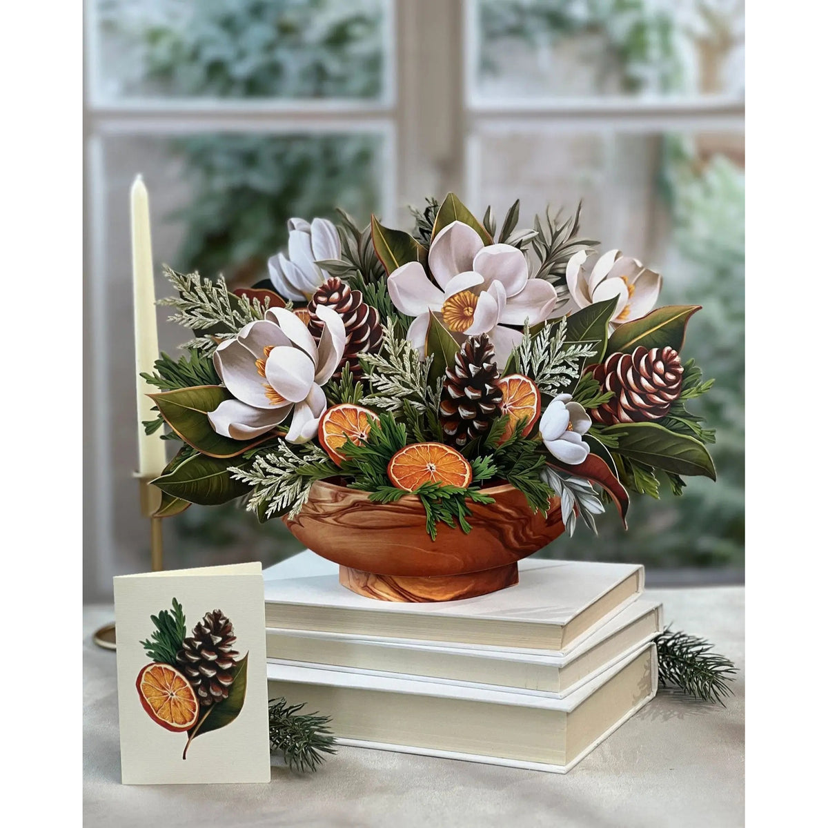 Decorative floral arrangement with oranges, pinecones, and a candle on books.