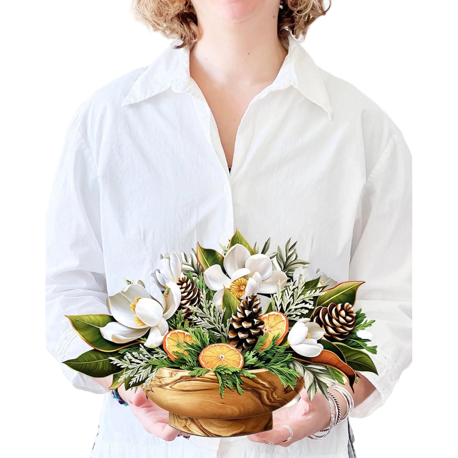 Person holding a wooden bowl with a floral arrangement featuring white flowers, green leaves, and pinecones.