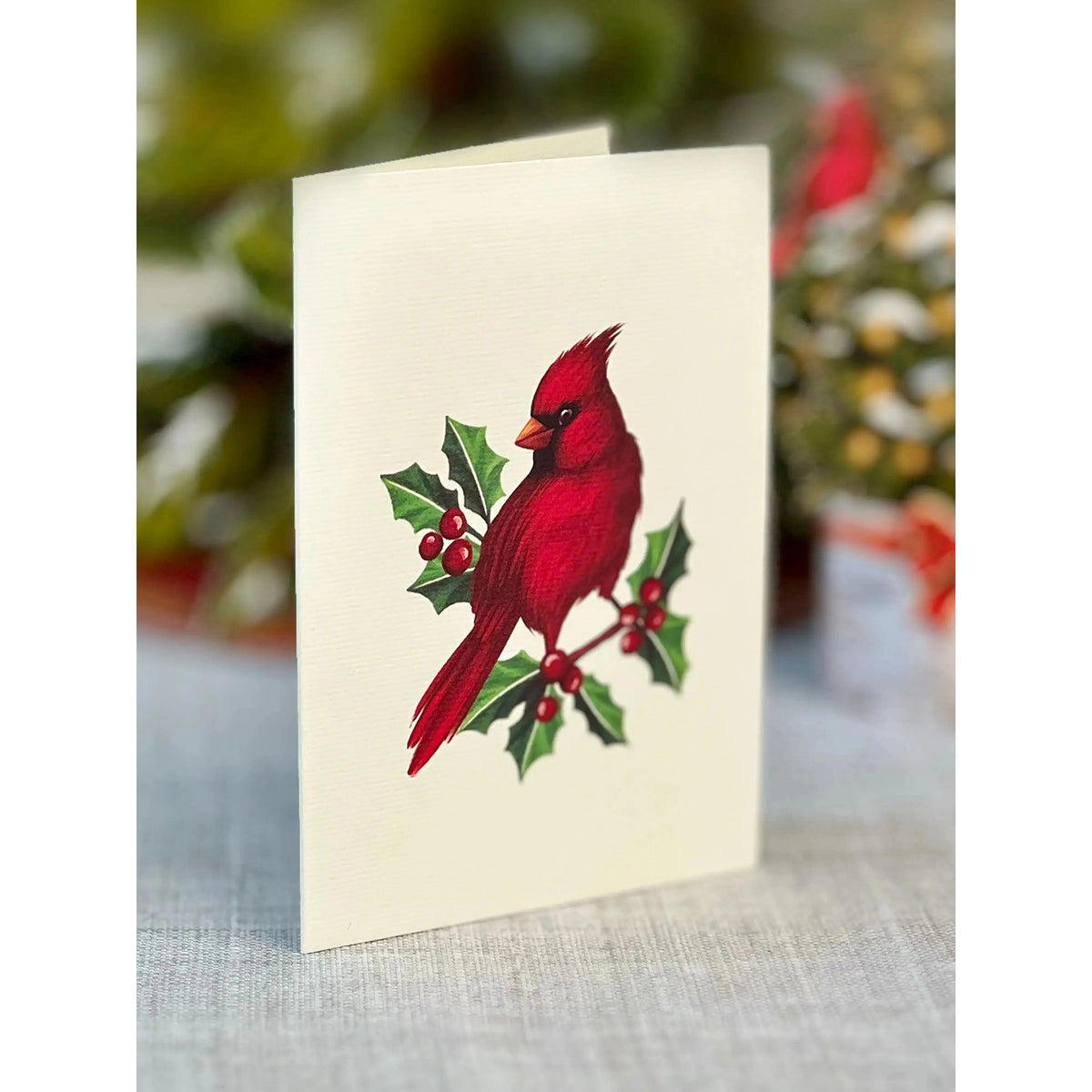 Card with a red cardinal bird and holly leaves on a textured surface with blurred Christmas decorations in the background.