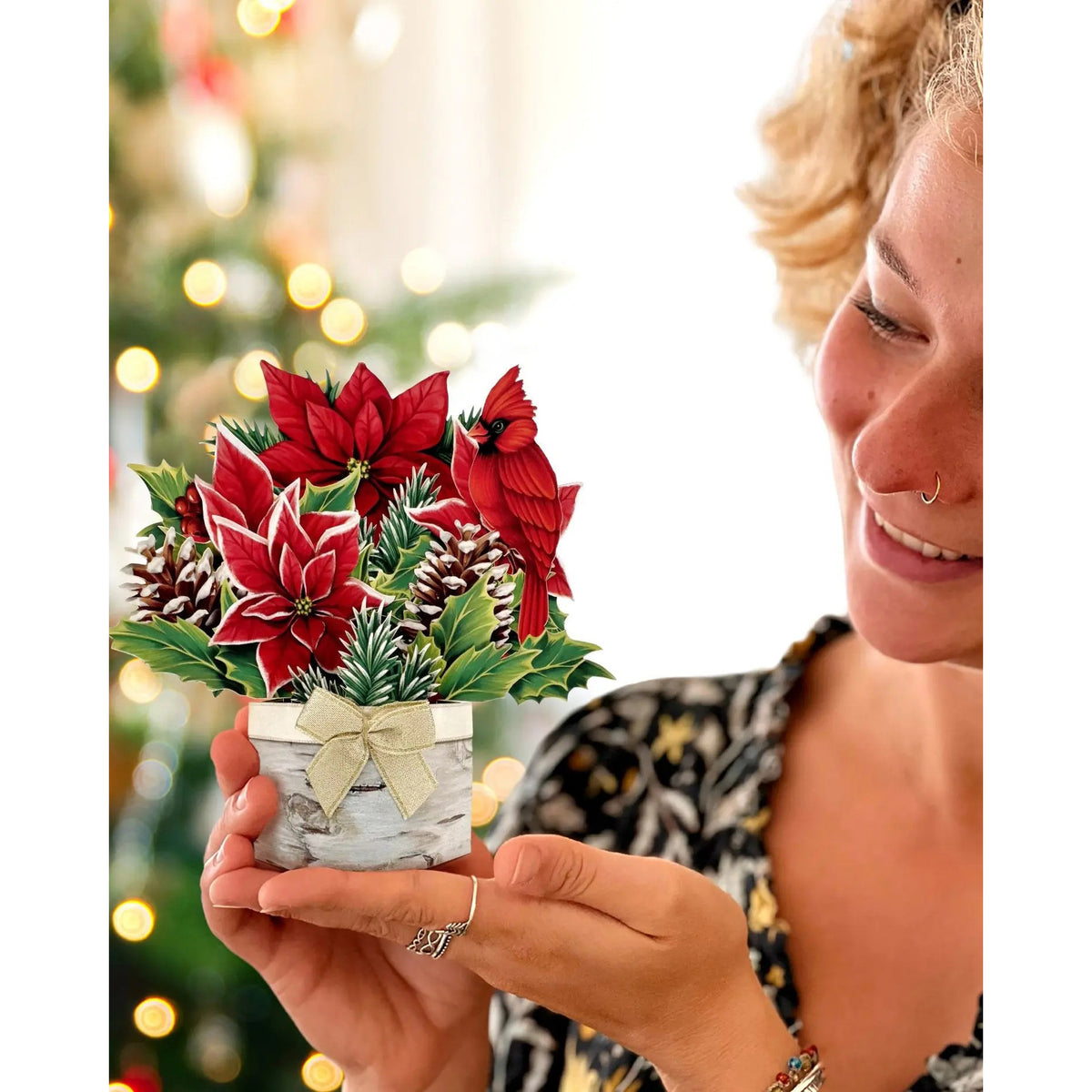 Person holding a small potted plant with red flowers and greenery, blurred Christmas tree in the background