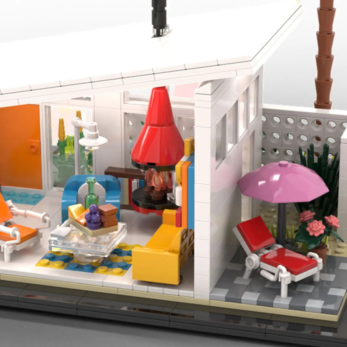 A detailed brick-built model of a modernist house interior, featuring a red fireplace, yellow and blue sofas, a pink patio umbrella, and red lounge chairs on a gray tiled deck.