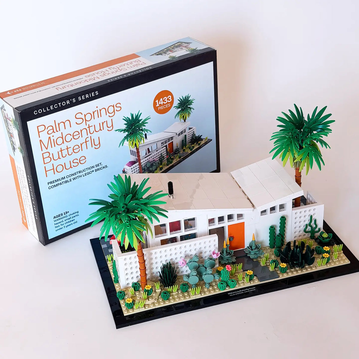 A boxed building-block set labeled 'Palm Springs Midcentury Butterfly House' sits behind a completed 3D model of the house. The model features white mid-century architecture with an orange door, green palm trees, cacti, and a landscaped front yard on a black base, photographed on a clean white background.