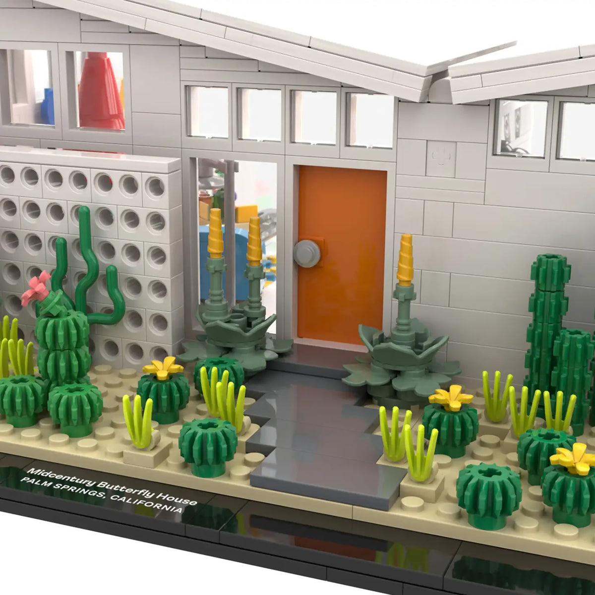 A LEGO-style mid-century modern butterfly house set with a gray brick exterior, orange door, and a landscaped garden with green cacti, yellow flowers, and gray stepping stones on a display base.