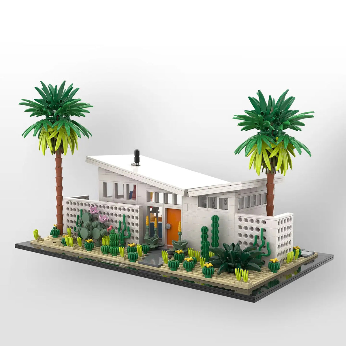 A modernist-style toy house model with two palm trees, cacti, and desert plants, displayed on a black base against a light gray background.
