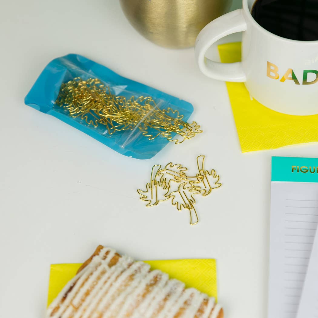 Gold metal paperclip charms shaped like leaves or pinecones, shown with a small blue pouch and loose pieces on a white desk alongside a coffee mug, notepad, and pastry.