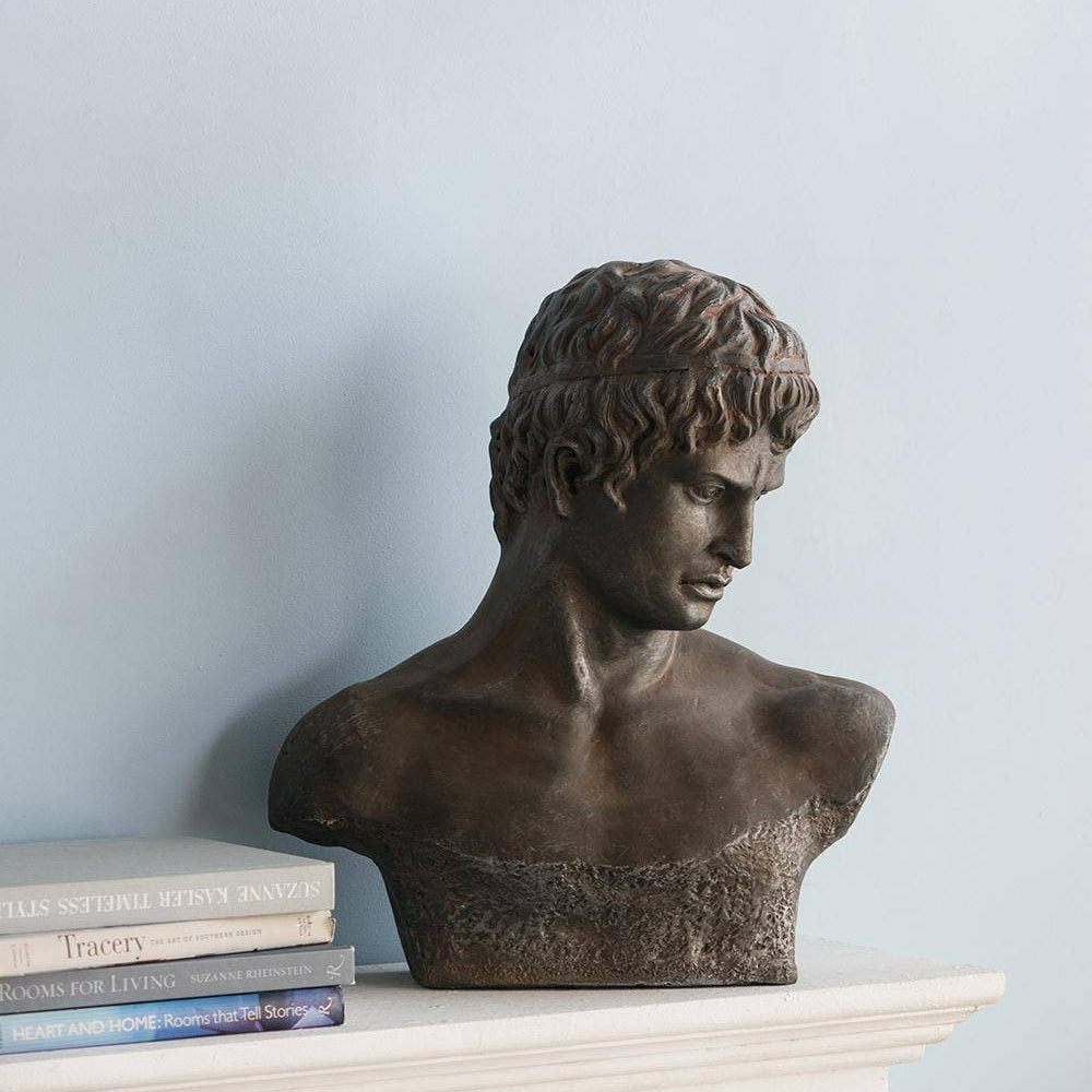 A dark bronze classical male bust sculpture displayed on a white mantelpiece next to a stack of books against a light blue wall.