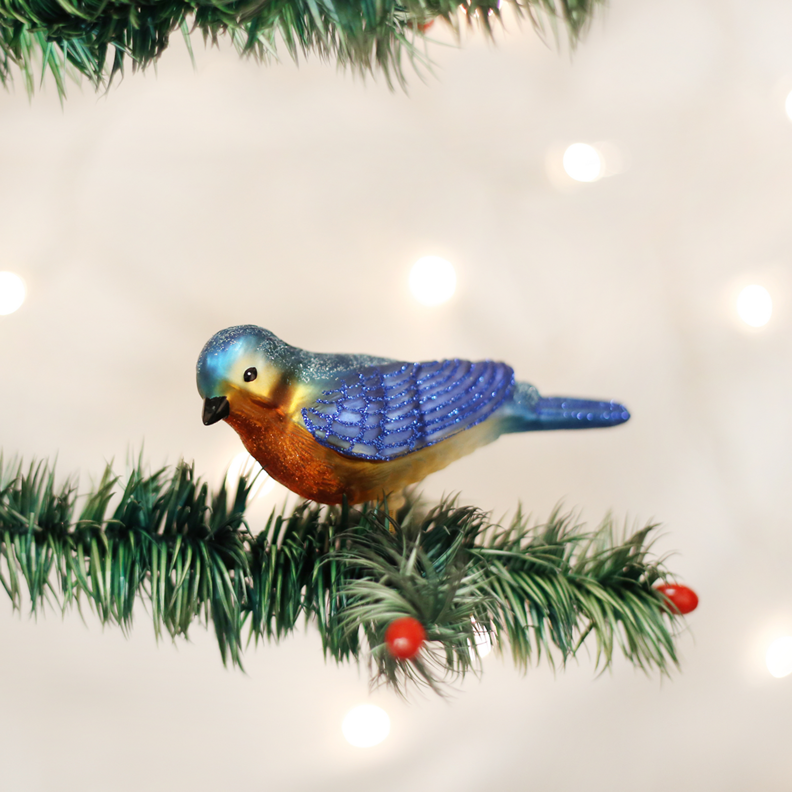 A decorative bird ornament shaped like a bluebird, with a glittery blue head and wings, a warm orange chest, and a gold metal clip at the bottom, shown on a plain white background.