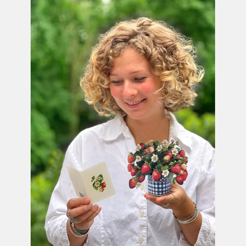 Woman holding a small potted plant and a card with floral design outdoors.