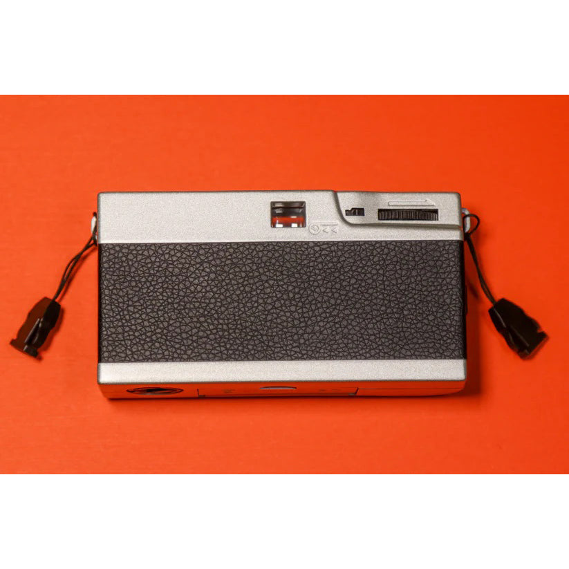 A compact 35mm film camera with a matte silver finish and black leatherette center, placed on an orange background.