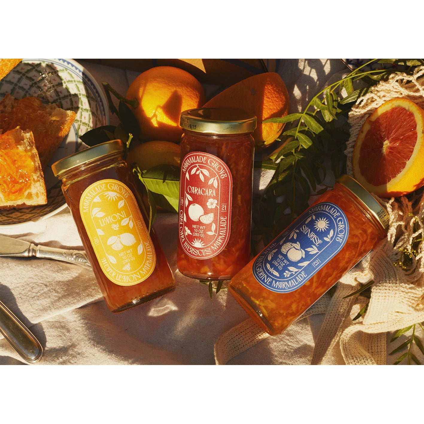 Three jars of marmalade n a box with oranges and bread on a table.