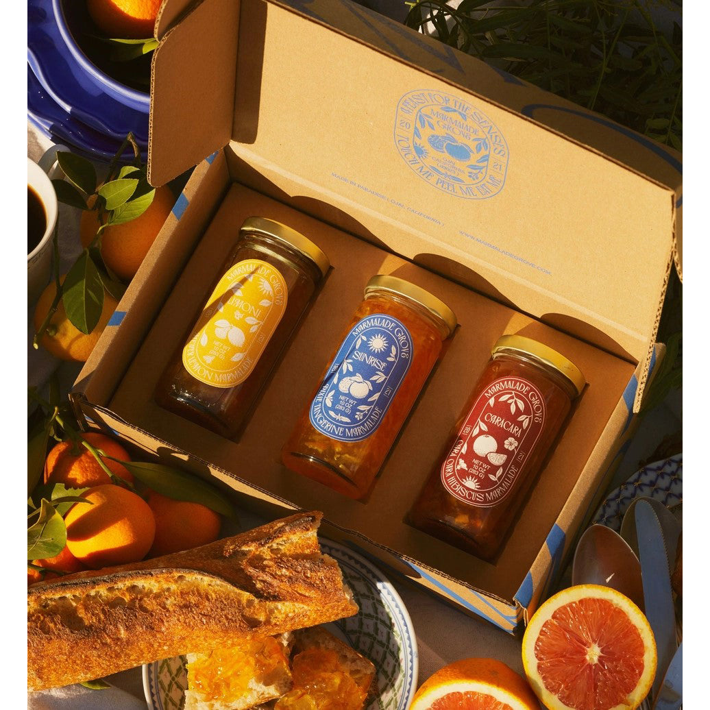 Three jars of marmalade n a box with oranges and bread on a table.