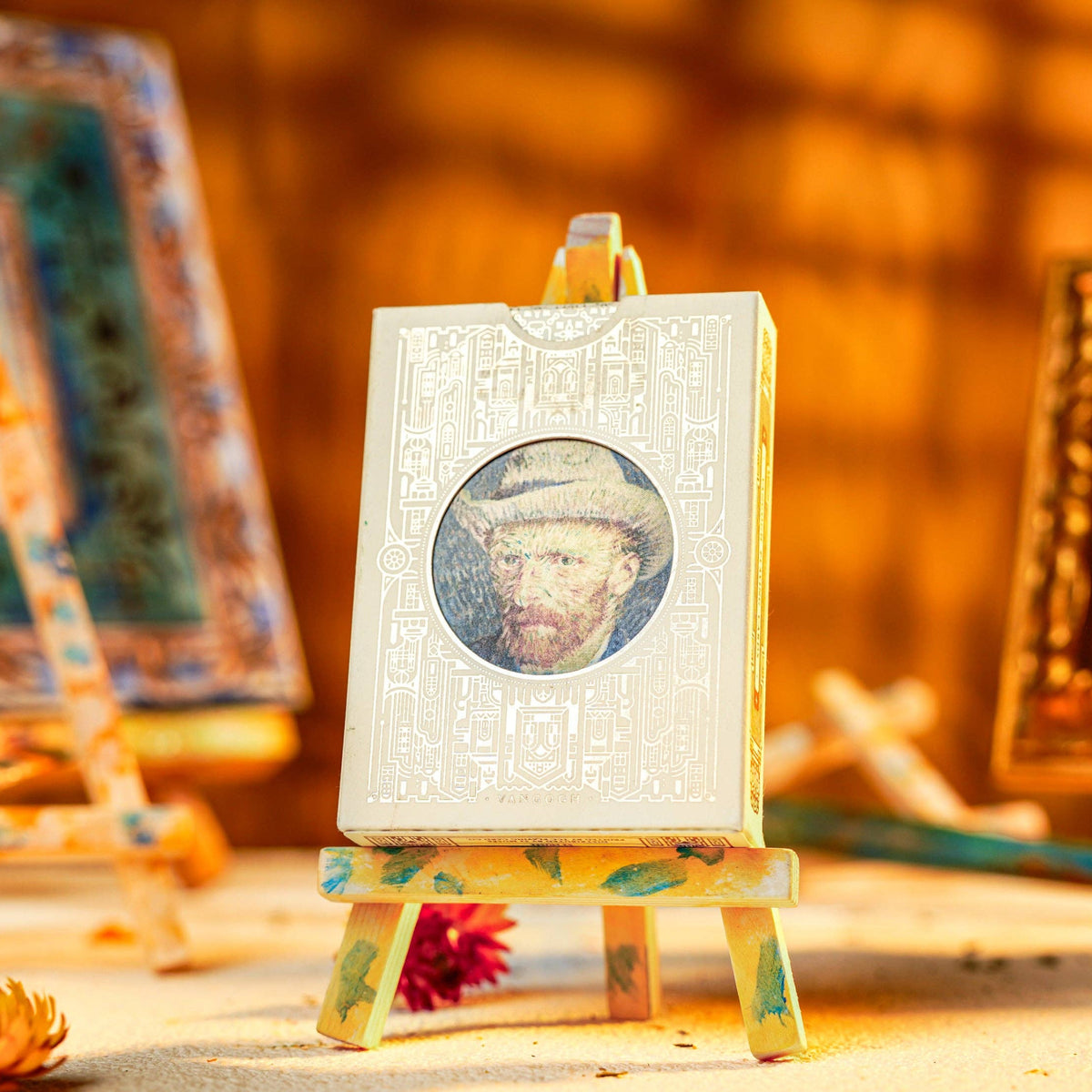 A miniature white canvas print on a small wooden easel, featuring a circular portrait. The scene is styled with warm, golden lighting, surrounded by other miniature art frames, canvases, and paint tubes.