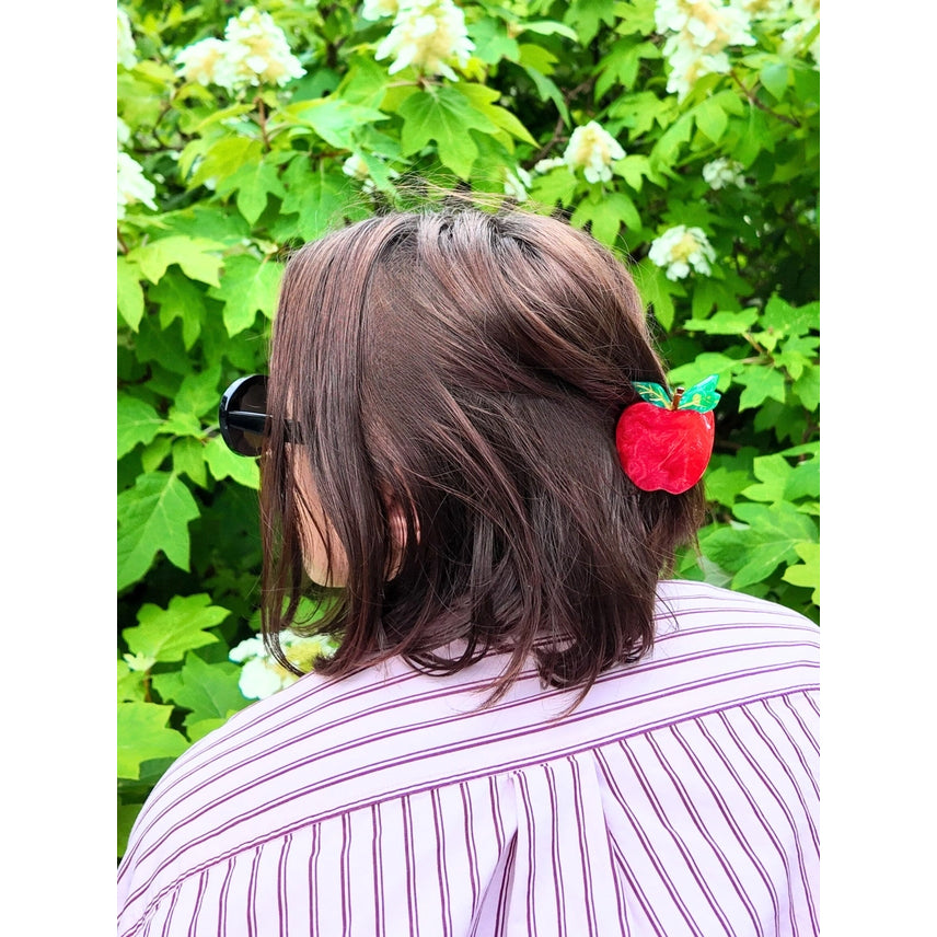 Person with a red hair clip shaped like an apple against a green leafy background
