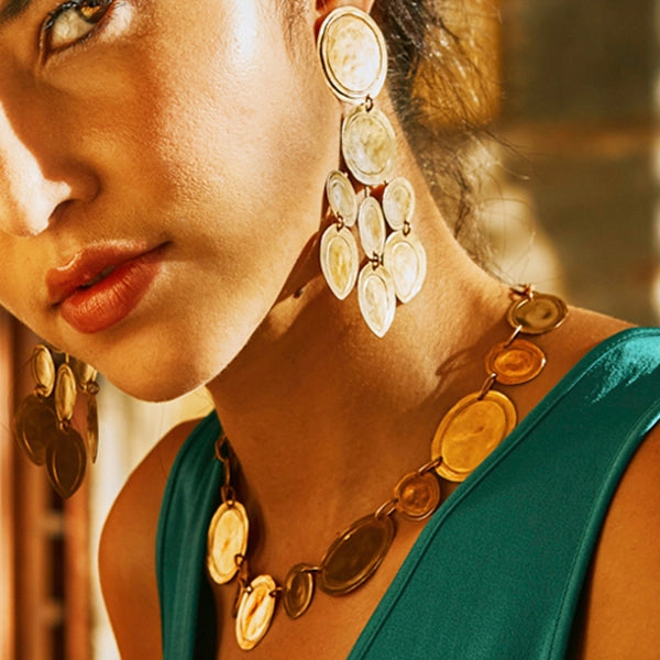A close-up lifestyle shot of a woman wearing chunky gold coin-link statement necklaces and matching drop earrings with oval cream and gold charms, paired with a teal green top.