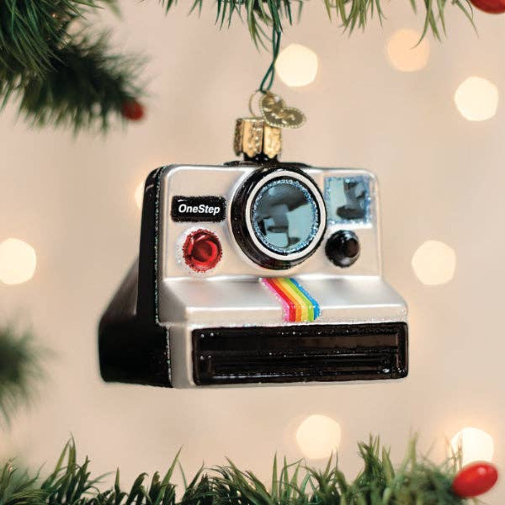 A Polaroid OneStep camera-shaped Christmas ornament hanging from a green tree branch, with warm bokeh lights in the background.