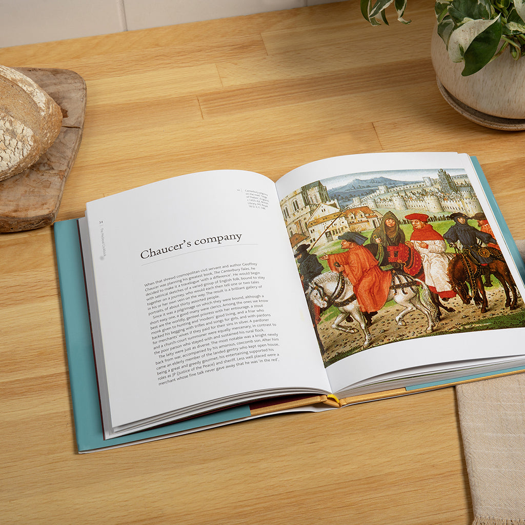 An open hardcover book lying on a light wood tabletop, showing a printed page with text and a large reproduction of a historical scene with people on horseback. The book cover edge is teal, and nearby props include a loaf of bread, a potted plant, and a folded cloth.