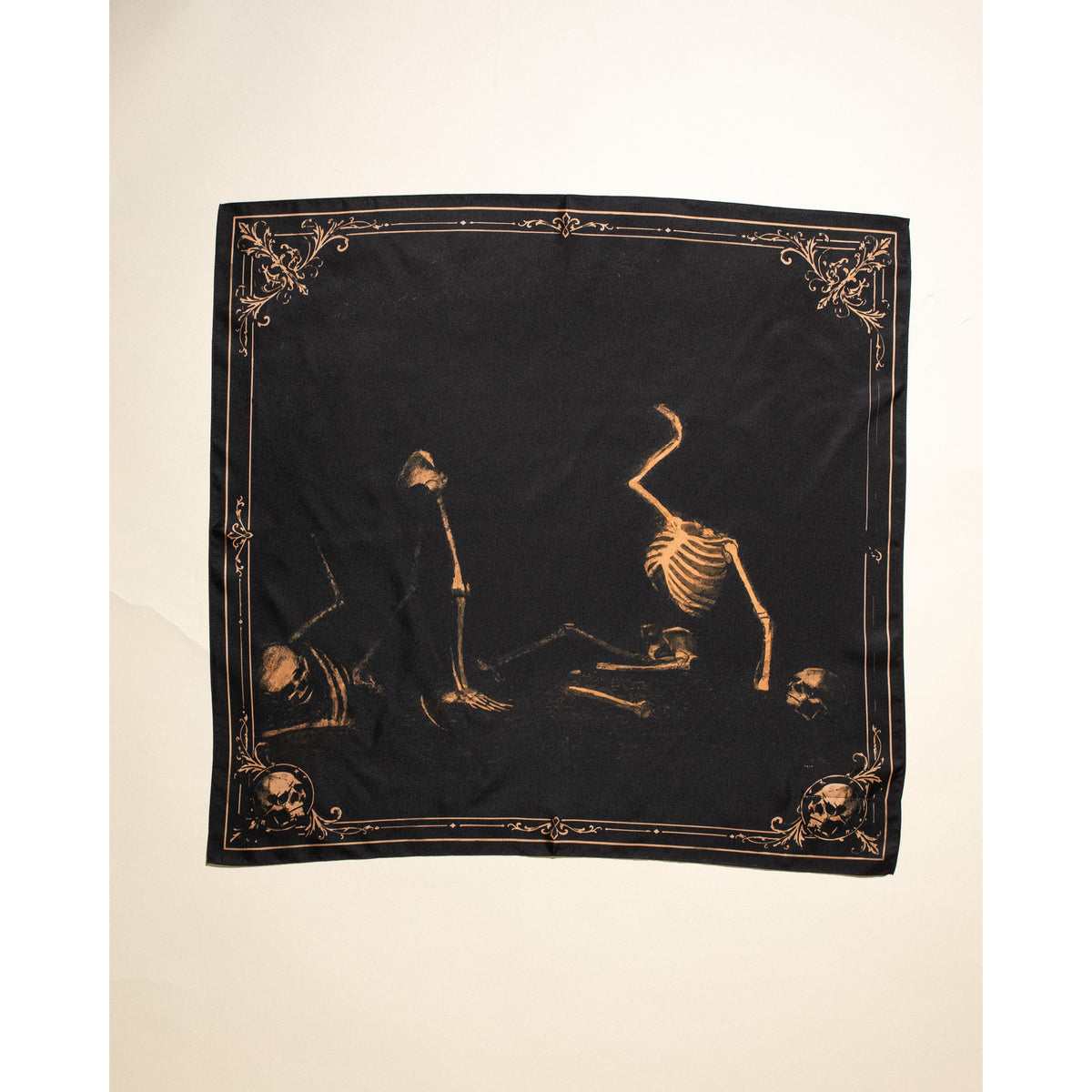 A square black scarf featuring a gothic design with three skeleton figures and ornate gold corner flourishes, displayed flat against a light beige background.