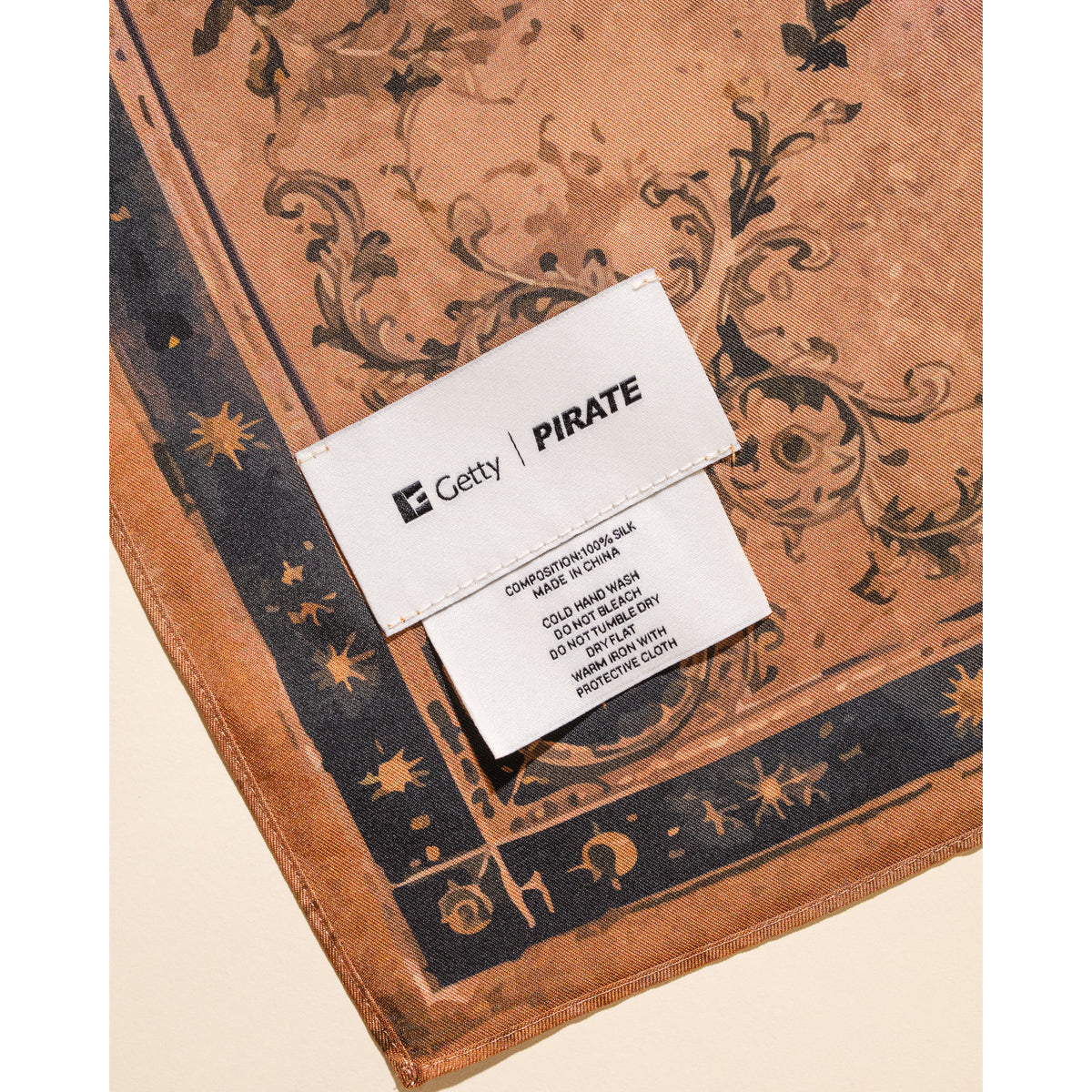 A close-up of a pirate-themed silk scarf with a brown and black baroque pattern, featuring a white paper label sewn onto the corner with 'Getty | PIRATE' printed on it.