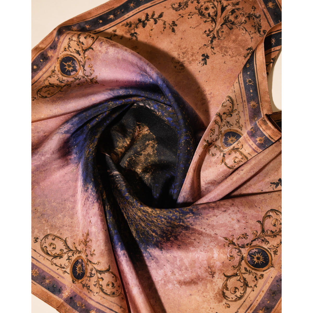 A square silk scarf with a faded bronze and dusty pink background, featuring dark blue borders, ornate baroque scrollwork, and a central dark blue and gold abstract pattern, draped in a spiral.